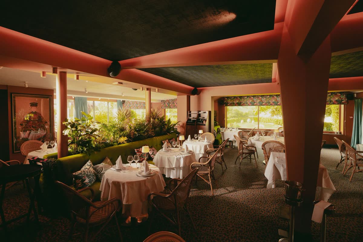 De zaal van Le Barbizon gedekt voor een diner: ronde tafels in wit linnen, rieten stoelen en een groene zitbank